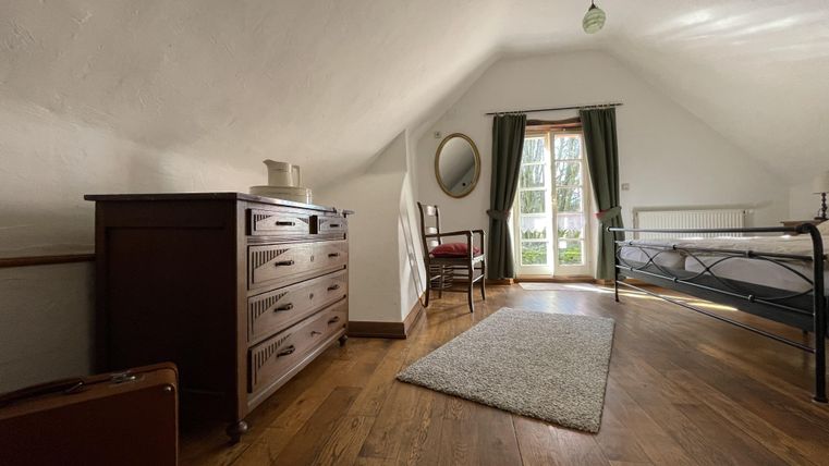 Une chambre confortable sous les combles avec un sol en bois et deux fenêtres. Il y a un lit, une commode et une chaise, qui créent une atmosphère accueillante.