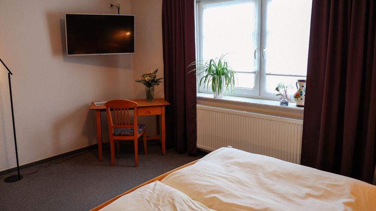 Une chambre simple avec un lit, une table et une télévision. Les fenêtres laissent entrer beaucoup de lumière du jour et il y a une plante sur la table.
