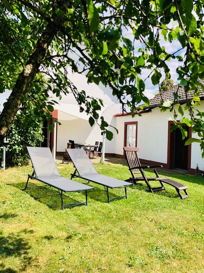 A quiet garden with two loungers and a chair. In the background, a bright house can be seen, surrounded by trees.