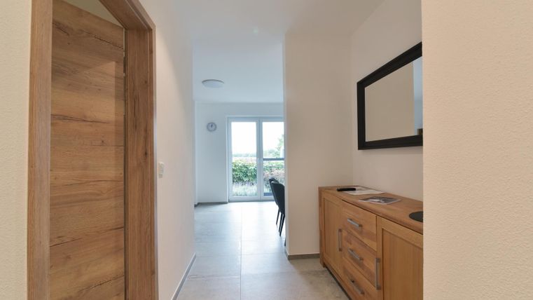 Een lichte gang met houten details en een moderne spiegelwand. Op de achtergrond zijn ramen die veel licht binnenlaten.