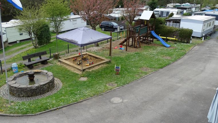 Un camping avec aire de jeux, bac à sable et toboggan. Dans les environs, il y a des camping-cars et des arbres.
