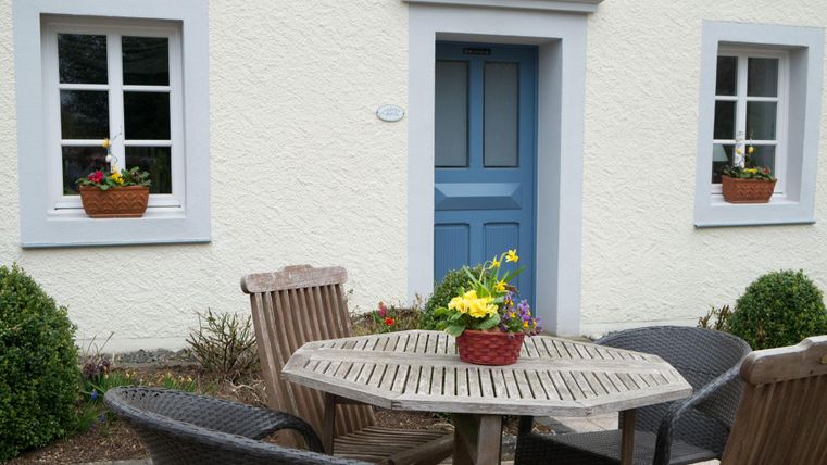 Een gezellige buitenruimte met een ronde houten tafel en stoelen. De ramen zijn versierd met bloembakken en een blauw gelakte deur trekt de aandacht.