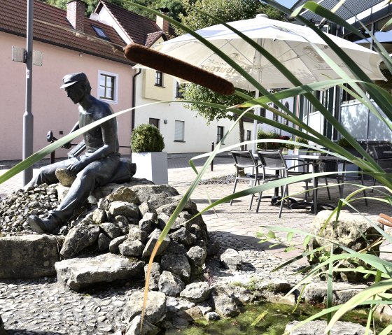 Bronzestatue eines Mannes auf Steinen, umgeben von Schilf und einem kleinen Teich. Im Hintergrund sind Häuser und ein Sonnenschirm zu sehen., © Tourist-Information Bitburger Land