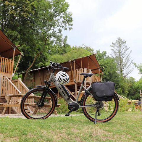 Ein modernes E-Bike steht auf einer Wiese vor mehreren Holzhütten. Im Hintergrund sind grüne Bäume und ein bewölkter Himmel zu sehen.
