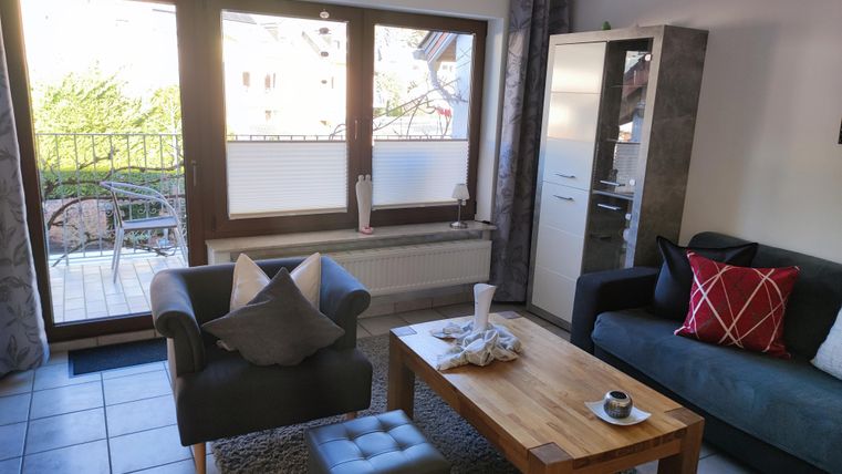 Ein gemütliches Wohnzimmer mit einem Sofa, Sesseln und einem Holztisch. Große Fenster bieten einen Blick auf einen Garten.