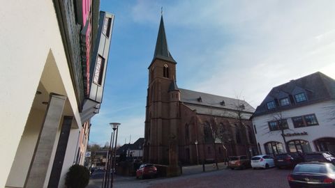 Eine historische Kirche mit einem spitzen Turm steht in einer Stadtstraße. Umgeben von modernen Gebäuden und Autos ist der Himmel klar und blau.
