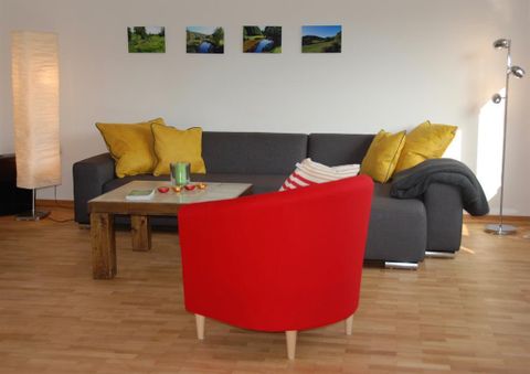 Ein gemütliches Wohnzimmer mit einer grauen Couch und gelben Kissen. Ein roter Stuhl und ein Holztisch vervollständigen die einladende Atmosphäre.