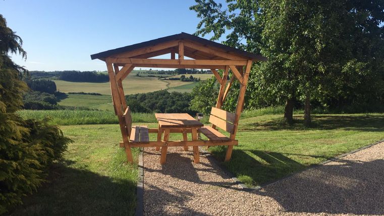 A covered outdoor seating area in front of the holiday home with a view. In the background, gentle hills and a clear blue sky can be seen.