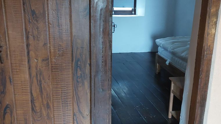 Een houten raam met een open deur leidt naar een lichte kamer. Op de achtergrond is een bed en een raam te zien.