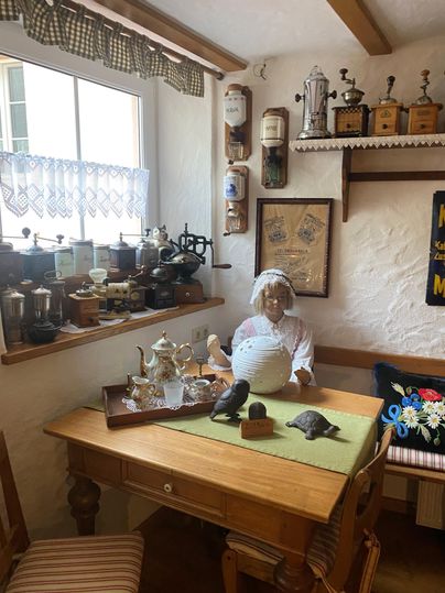 Une pièce confortable avec une table et divers objets décoratifs. Une poupée en vêtements traditionnels est assise dans le coin et confère à la pièce un charme nostalgique.