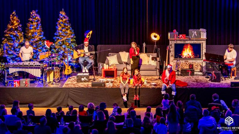 Eine festliche Weihnachtsaufführung mit Musikern auf der Bühne. Im Hintergrund stehen Weihnachtsbäume und ein Feuerplatz sorgt für eine gemütliche Atmosphäre.