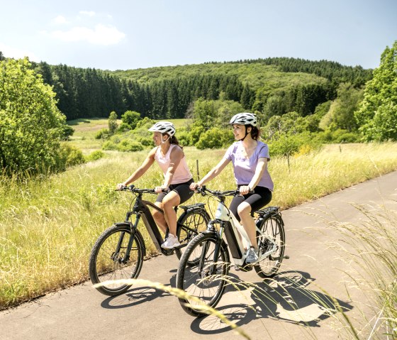 Zwei Radfahrer mit Helmen fahren auf einem asphaltierten Weg durch eine gr&uuml;ne, bewaldete Landschaft unter blauem Himmel., &copy; Eifel Tourismus GmbH, Dominik Ketz