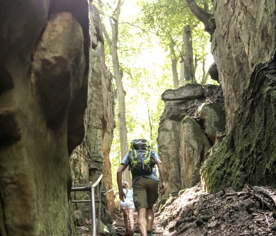 Zwei Personen wandern durch die Teufelsschlucht, umgeben von hohen Felsen und Bäumen. Sonnenlicht strahlt durch das Blätterdach., © Eifel Tourismus GmbH, Dominik Ketz