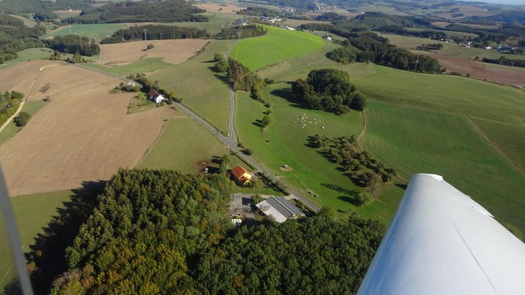 Een luchtfoto van een landelijke landschap met velden en bossen. In de afbeelding zijn enkele gebouwen en een weg zichtbaar die zich door de groene omgeving slingert.