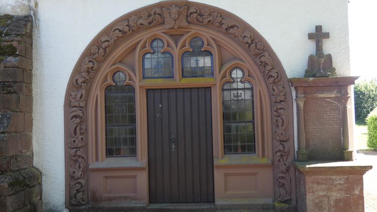 Ein historischer Gebäudeeingang mit aufwendigen Verzierungen. Das Tor ist dunkelbraun und wird von dekorativen Fenstern flankiert.