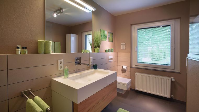 Une salle de bain moderne avec des murs clairs et un grand lavabo. La décoration verte et beaucoup de lumière naturelle donnent à l'espace une atmosphère fraîche.