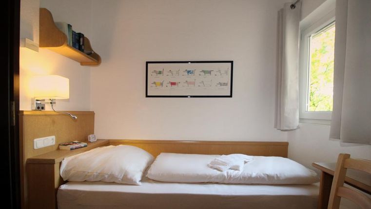 Ein einfaches Zimmer mit einem Einzelbett und einem Schreibtisch. Das Zimmer hat ein Fenster mit Tageslicht und dekorative Bilder an der Wand.