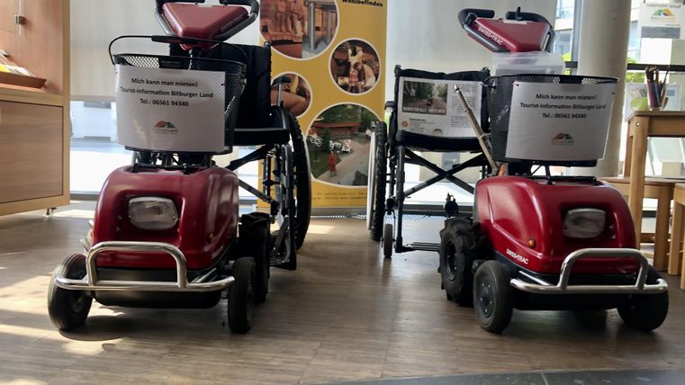 Twee rode gemotoriseerde rolstoelen staan naast elkaar in een binnenruimte. Op de achtergrond zijn informatieschermen te zien.
