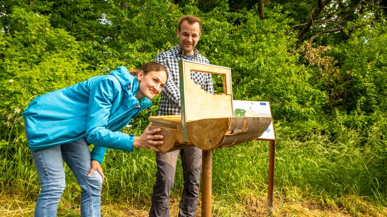 Zwei Personen betrachten ein hölzernes Objekt in einer grünen Umgebung.