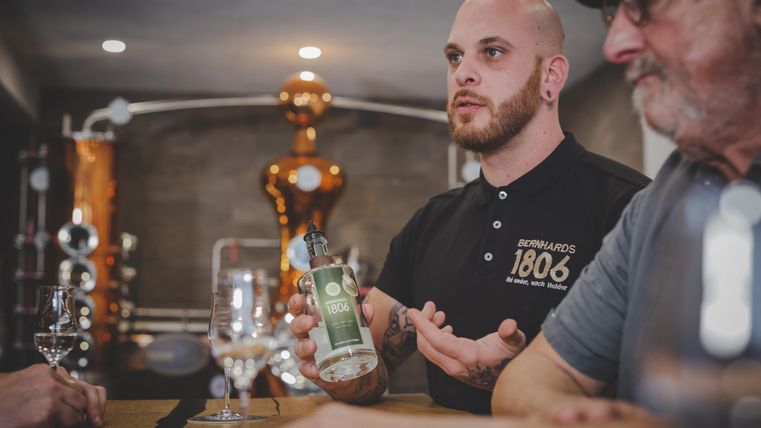 Ein junger Mann erklärt etwas an einer Bar mit einem Getränk in der Hand. Im Hintergrund sind Destilliergeräte sichtbar.
