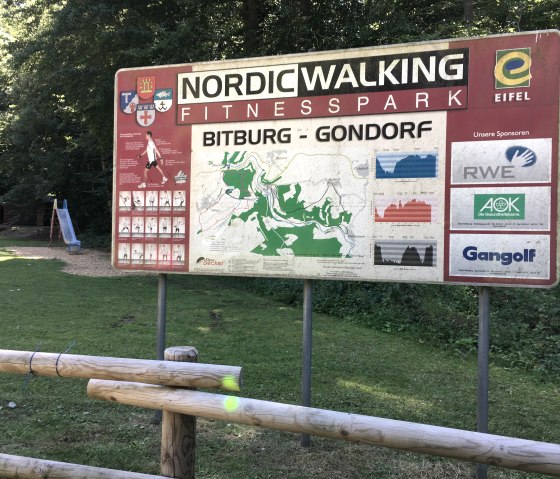 Sign for the Bitburg-Gondorf Nordic Walking Fitness Park with map and sponsor logos in the forest, next to a playground with slide., &copy; TI Bitburger Land
