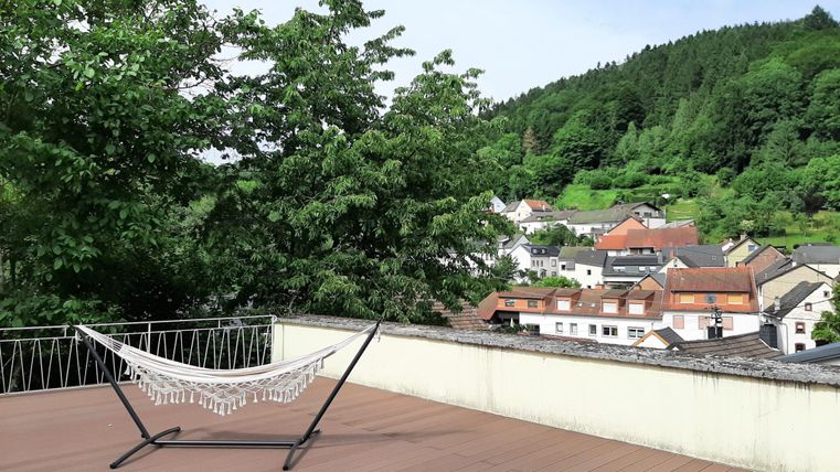 Eine helle Terrasse mit einer Hängematte und Blick auf das Dorf und die umliegenden Bäume. Die Landschaft ist grün und einladend.