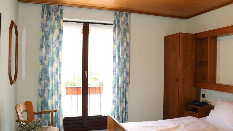 Une chambre simple avec un lit, une chaise et une table. De grandes fenêtres avec des rideaux laissent entrer beaucoup de lumière.