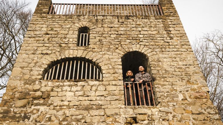 Zwei Personen stehen auf einem Balkon einer alten Steinburg mit Gitterfenstern.