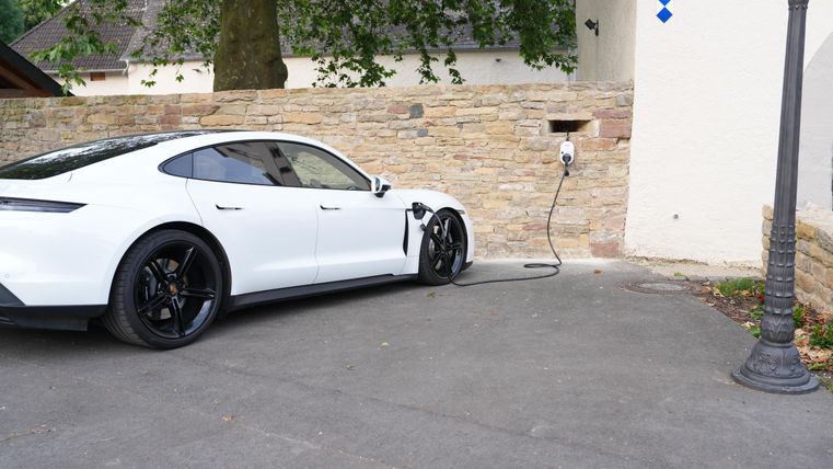 Ein weißes Elektroauto steht an einer Ladesäule. Im Hintergrund ist eine Steinmauer und ein Laternenpfahl sichtbar.