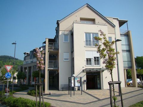 Ein modernes Gebäude mit mehreren Etagen und großen Fenstern. Umgeben von Bäumen und einem gepflegten Platz.