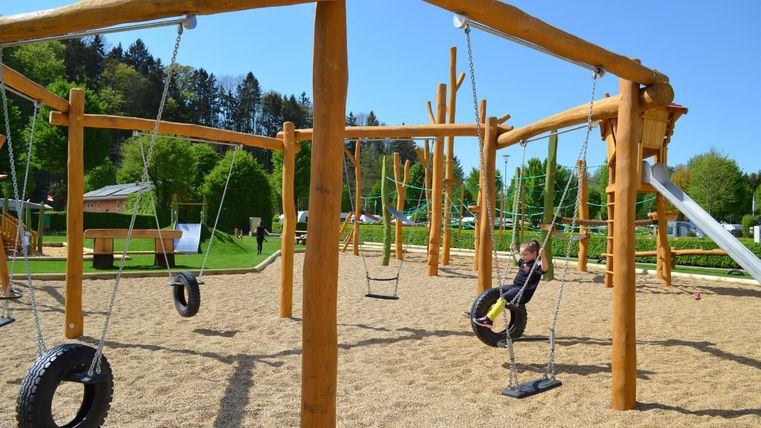 Ein schöner Spielplatz mit Holzspielgeräten und Schaukeln. Der Boden ist mit Sand bedeckt und es ist sonnig.
