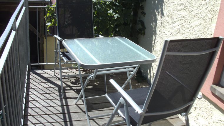 Ein kleiner Balkon mit einem Tisch und zwei Stühlen. Die Oberfläche ist aus Holz und es gibt Pflanzen in der Nähe.