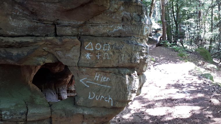 Une formation rocheuse dans la forêt avec des symboles et des flèches illisibles. Le chemin traverse un environnement verdoyant.