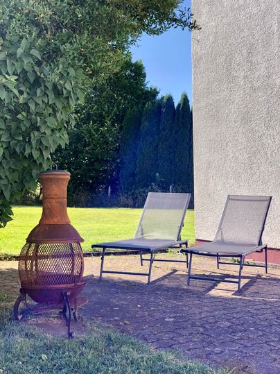 A cozy outdoor area with two loungers and a rustic fireplace. The green lawn and the blue sky create a relaxed atmosphere.