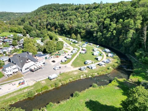 Een camping naast een rivier, omringd door bomen en heuvels. Meerdere campers en tenten zijn verspreid over het terrein.