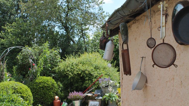 Eine rustikale Gartenansicht mit hängenden Kochutensilien an einer Wand. Im Hintergrund sind üppige Pflanzen und Blumen zu sehen.