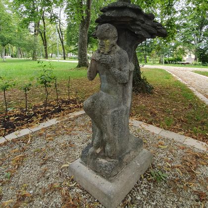 Une statue d'un jeune homme assis tenant un objet dans ses mains. Autour de lui, il y a des arbres et une zone de jardin.