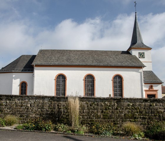 Die Pfarrkirche St. Martin in Dockendorf mit Uhrturm, umgeben von einer Steinmauer und einem Garten. Der Himmel ist leicht bew&ouml;lkt., &copy; TI Bitburger Land