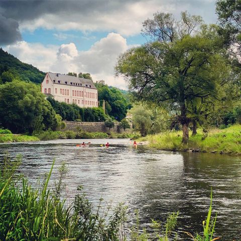Ein ruhiger Fluss mit Kajakfahrern und üppigen Bäumen am Ufer. Im Hintergrund steht ein historisches Gebäude unter einem bewölkten Himmel.