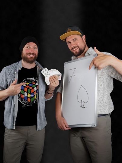 Zwei Männer stehen vor einem dunklen Hintergrund. Einer hält eine Spielkarte, während der andere ein Zeichen mit einer Pik-Sieben präsentiert.
