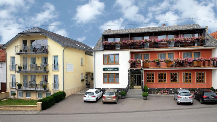 Ein kleines Hotel mit zwei Gebäuden und Balkonen. Vor dem Hotel stehen einige Autos und der Himmel ist teilweise bewölkt.