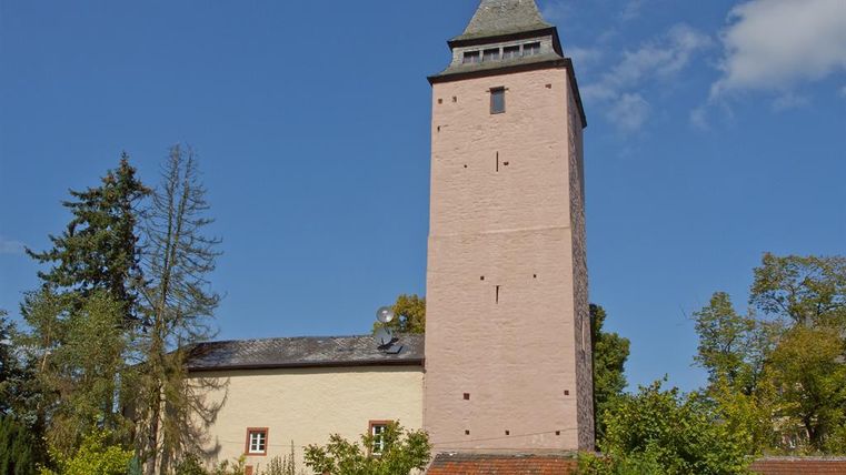 Ein historischer Turm umgeben von grünen Bäumen und Sträuchern. Der Himmel ist klar und blau.