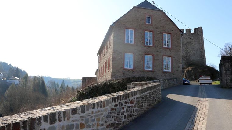 Ein altes Steingebäude mit Fensterläden und einem historischen Charakter. Die Straße führt sanft an der Fassade vorbei.