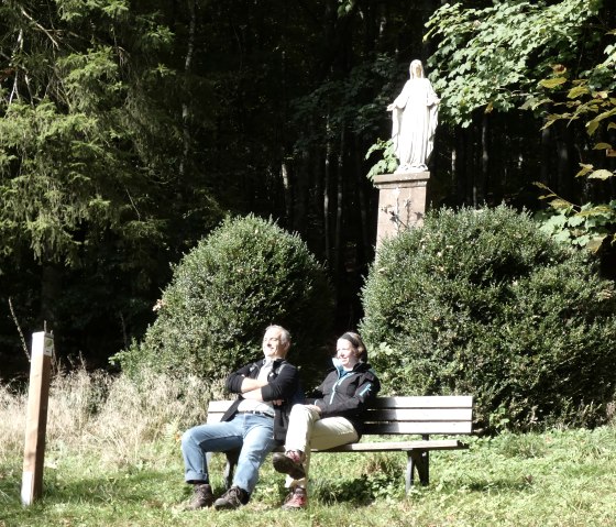 Zwei Personen sitzen auf einer Bank vor einer Mariens&auml;ule im Wald. Sie genie&szlig;en die Sonne, umgeben von B&uuml;schen und B&auml;umen., &copy; TI Bitburger Land