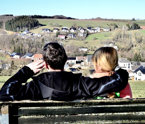 Ein Paar sitzt auf einer Bank und schaut auf das Dorf Baustert in einer grünen, hügeligen Landschaft., © TI Bitburger Land