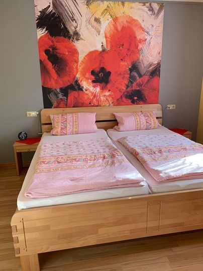 Een gezellige slaapkamer met een dubbelbed en roze bedloop. Op de achtergrond is een groot, helder schilderij met rode bloemen te zien.