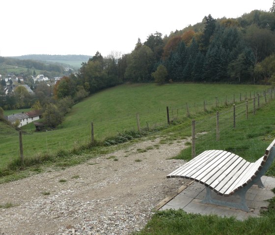Eine geschwungene Holzbank steht auf einem Hügel mit Blick auf Mettendorf, umgeben von grünen Wiesen und Bäumen., © Felsenland Südeifel Tourismus GmbH, Christian Calonec-Rauchfuss