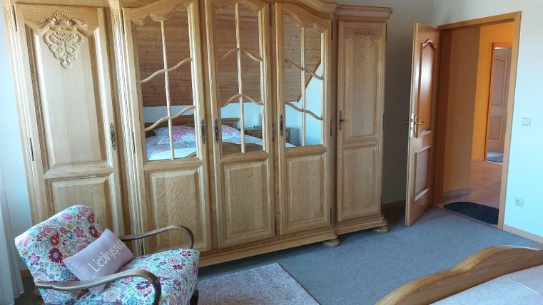 Een gezellige slaapkamer met een grote houten kast en een bloeiende stoel. De muren zijn licht geverfd en er is toegang tot een andere kamer.