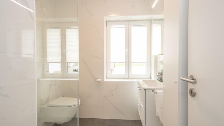 Une salle de bain moderne avec une douche en verre et des murs clairs. De grandes fenêtres laissent entrer beaucoup de lumière du jour.