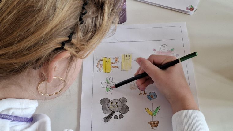 Ein Kind malt mit einem grünen Stift auf einem Blatt Papier. Die Zeichnung zeigt ein Elefant und verschiedene farbige Motive.